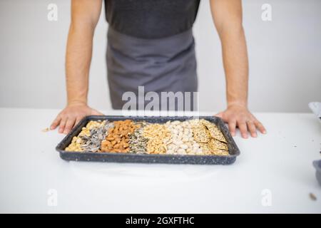 Man showing tray of chickpeas, almonds, peanuts, pistachios, and crispbread. Fresh crispy bread, seeds, and nuts on white table. Healthy snacks Stock Photo
