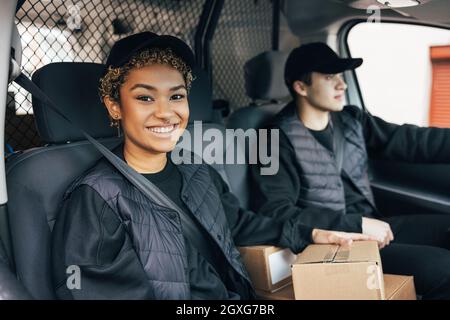 Smiling woman in uniform sitting in delivery van and looking at camera while her coworker driving Stock Photo