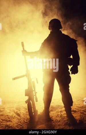 Army sniper with large caliber rifle standing in the fire and smoke. Backlit silhouette, toned image Stock Photo