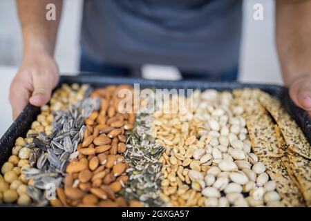 Male hands holding black tray of chickpeas, almonds, peanuts, pistachios, and multi-seed crispbread. Fresh crispy bread, seeds, and nuts. Healthy snac Stock Photo