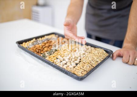 Man showing tray of chickpeas, almonds, peanuts, pistachios, and crispbread. Fresh crispy bread, seeds, and nuts on white table. Healthy snacks Stock Photo