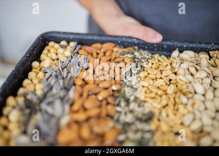 Male hands holding black tray of chickpeas, almonds, peanuts, pistachios, and multi-seed crispbread. Fresh crispy bread, seeds, and nuts. Healthy snac Stock Photo