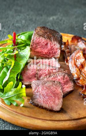 Sliced beef steak with fried rashers of bacon and leafs of fresh arugula on the wooden board Stock Photo
