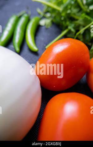 Top view of three green chili peppers, onion, four tomatoes, and coriander on table. Fresh and aromatic vegetables and herbs on dark blue surface. Hea Stock Photo