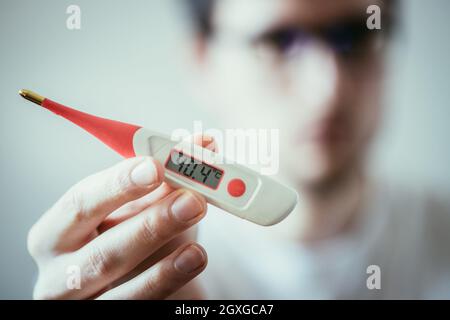 Man holds a red fever thermometer with high temperature in his hand, blurry face Stock Photo