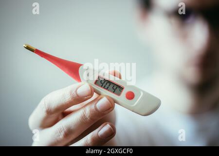 Man holds a red fever thermometer with high temperature in his hand, blurry face Stock Photo