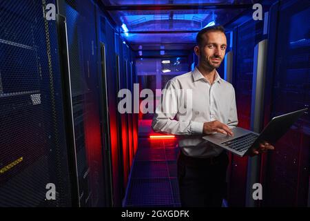Bearded smiling it engineer in white shirt working with server rack and telecommunication equipments in data center Stock Photo