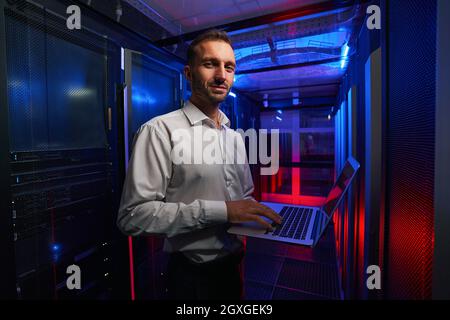 Bearded smiling system administrator is working with server rack and telecommunication equipments in data center Stock Photo
