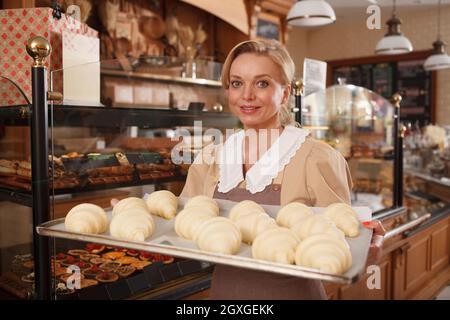 Lovely female baker smiling joyfully, carrying raw croissants to baking oven at her pastry shop Stock Photo
