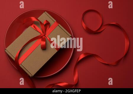 rectangular brown cardboard box tied with a silk red ribbon lies in a round ceramic red plate, top view Stock Photo