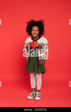 Cheerful african american girl in warm cardigan holding cup on red background Stock Photo