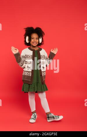 African american kid in ear muffs and cardigan pointing with hands on red background Stock Photo