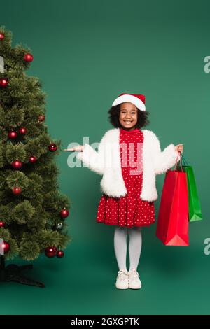 Happy african american girl in santa hat and fluffy jacket holding shopping bags and pointing at christmas tree on green background Stock Photo