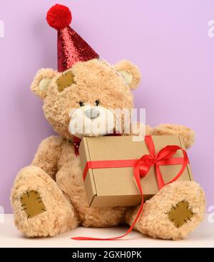 cute brown teddy bear in a red cap sits and holds a brown box with a gift, festive background Stock Photo