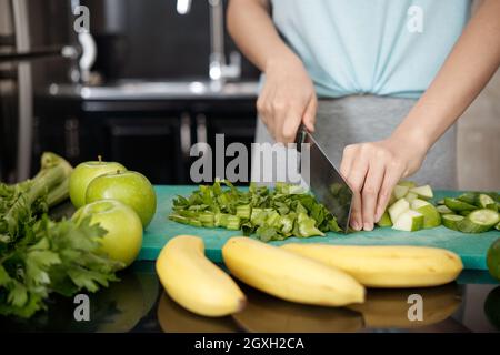 Close-up of unrecognizable woman standing at counter with vegetables and fruits and cutting celery on plastic board Stock Photo