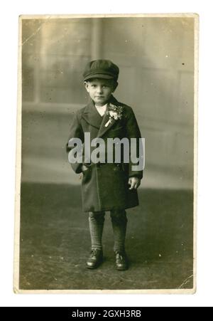 Original early 1940's postcard studio portrait of cute serious looking working class young lad wearing a smart long jacket, with buttonhole flowers on his lapel, and a flat cap. 1940's boy. 1940's child. 1940's children. He is maybe a pageboy at a wedding, Cheriton, Folkestone, Kent, U.K. dated July 6 1940 Stock Photo