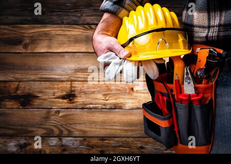 Electrician worker on vintage wooden background; holds helmet, gloves and goggles in hand. Construction industry, electrical system. Stock Photo