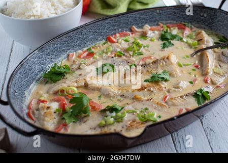 Fish dish with fried redfish fillet cooked in a delicious coconut milk with vegetables. Served in a rustic casserole dish on wooden table from above Stock Photo