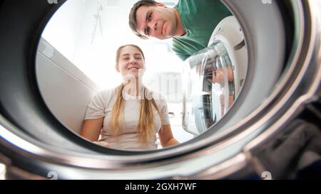 Smiling young couple looking inside washing machine and taking out clothes at laundry. Family doing housework and laundry together. Stock Photo