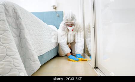 Housekeeper or maid in hotel wearing protective medical suit and mask washing and cleaning floor in room. Disinfection in hotels. Stock Photo