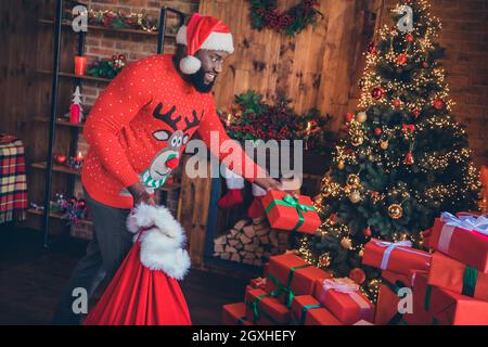 Photo of funny charming dark skin guy dressed print pullover smiling putting presents under christmas tree indoors house home room Stock Photo