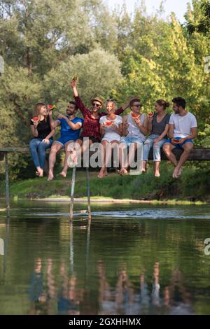group of young friends enjoying watermelon while sitting on the wooden bridge over the river in beautiful nature Stock Photo
