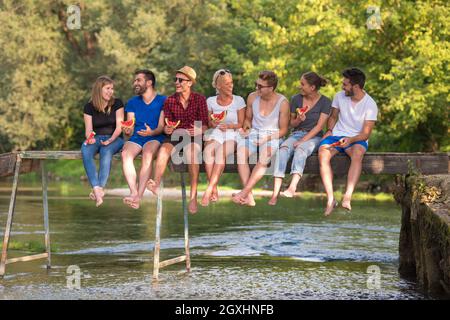 group of young friends enjoying watermelon while sitting on the wooden bridge over the river in beautiful nature Stock Photo