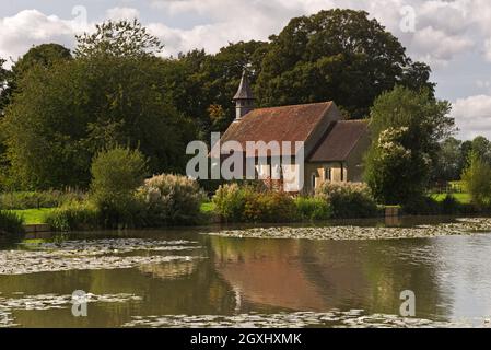 an idyllic view of St Leonards Church, Hartley Mauditt, Hampshire, England, UK with the still waters of the lake in the foreground. Stock Photo