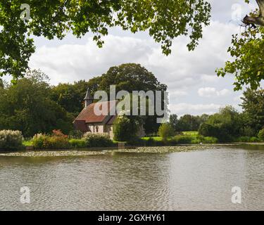 an idyllic view of St Leonards Church, Hartley Mauditt, Hampshire, England, UK with the still waters of the lake in the foreground. Stock Photo