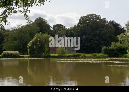 an idyllic view of St Leonards Church, Hartley Mauditt, Hampshire, England, UK with the still waters of the lake in the foreground. Stock Photo