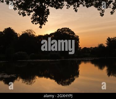 Sun setting behind St Leonards Church, Hartley Mauditt, Hampshire, England, UK with the still waters of the lake in the foreground. Stock Photo