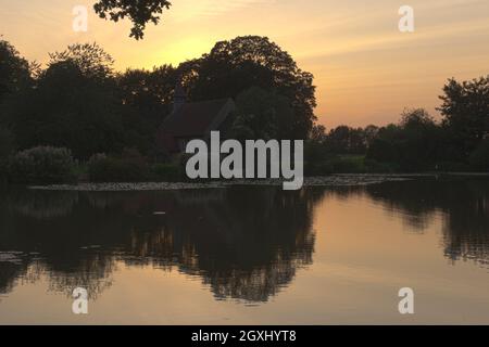 Sun setting behind St Leonards Church, Hartley Mauditt, Hampshire, England, UK with the still waters of the lake in the foreground. Stock Photo