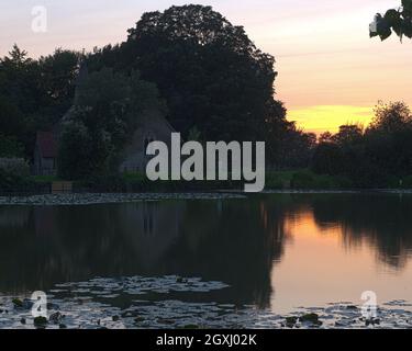 Sun setting behind St Leonards Church, Hartley Mauditt, Hampshire, England, UK with the still waters of the lake in the foreground. Stock Photo