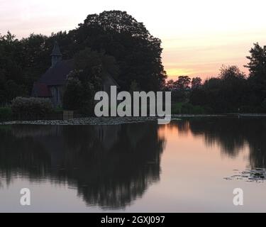 Sun setting behind St Leonards Church, Hartley Mauditt, Hampshire, England, UK with the still waters of the lake in the foreground. Stock Photo