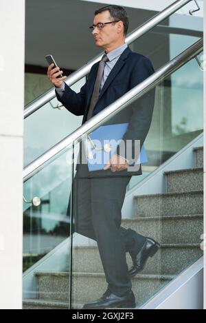 Serious mature Caucasian businessman in formal suit carrying folder and using smartphone while moving down stairs Stock Photo