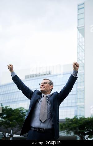 Excited mature Caucasian businessman in glasses and suit walking with raised arms while celebrating success outdoors Stock Photo