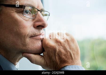 Headshot of thoughtful mature Caucasian man in glasses touching chin and contemplating cityscape through window Stock Photo
