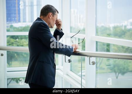 Tired mature businessman in suit standing at railings of office balcony and holding eyeglasses while massaging bridge of nose Stock Photo