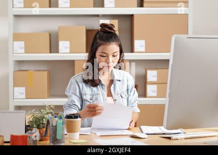 Busy young Asian woman with curly hair sitting at desk and working with documents Stock Photo