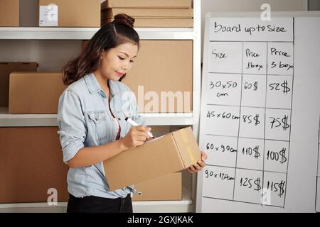 Serious young Asian postal worker standing against shelves with packages and signing parcel box Stock Photo