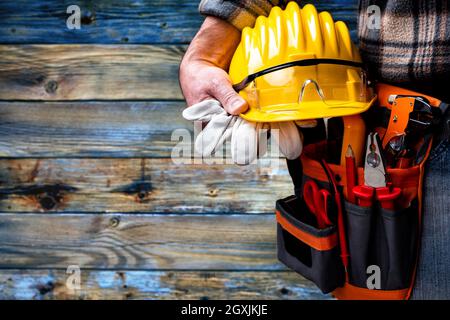 Electrician worker on vintage light blue wooden background; holds helmet, gloves and goggles in hand. Construction industry, electrical system. Stock Photo