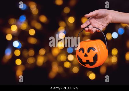 Children's hand holding an orange pumpkin-shaped basket, Jack's lantern on a dark background with beautiful bokeh. Waiting for Halloween candies. Tric Stock Photo