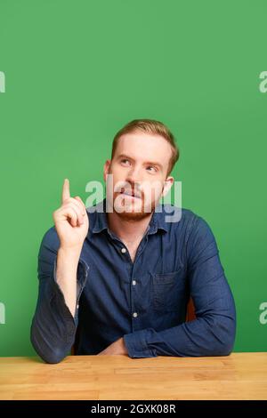 Pensive handsome redhead man in dark casual shirt sitting at wooden table and pointing finger up while thinking of idea Stock Photo