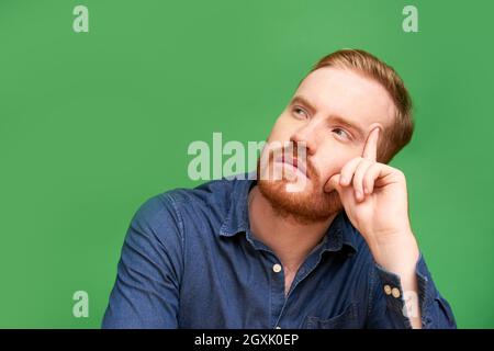 Serious redhead young man with beard sitting against green background and being deep in thoughts Stock Photo