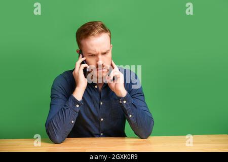 Puzzled young Caucasian man with beard sitting at wooden table against green background and talking on phone Stock Photo