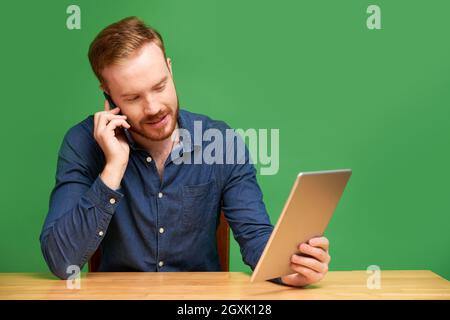 Smiling confident young Caucasian manager with beard sitting at desk and using tablet while talking by cellphone Stock Photo