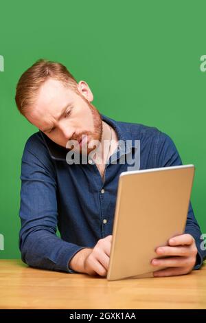 Concentrated young Caucasian man with beard sitting at wooden table and working with several devices, green background Stock Photo
