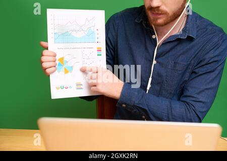 Close-up of fnancial consultant in earphones pointing at graph while explaining statistical data to client and talking to him via videoconferencing Stock Photo