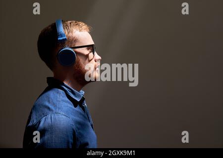 Side view of serious young red-bearded man listening to music in blue wireless headphones, dark room Stock Photo