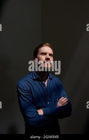 Serious pensive young bearded man in shirt standing with crossed arms in dark room Stock Photo